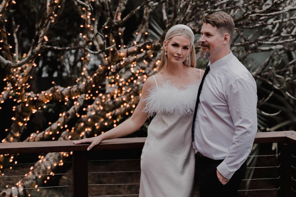 Couple poses together on wooden deck with twinkle lights creating romantic ambiance.