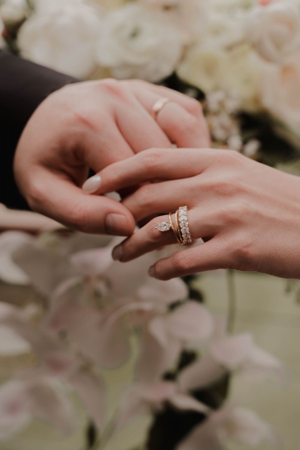 Close-up of hands exchanging diamond wedding rings with white florals in background.