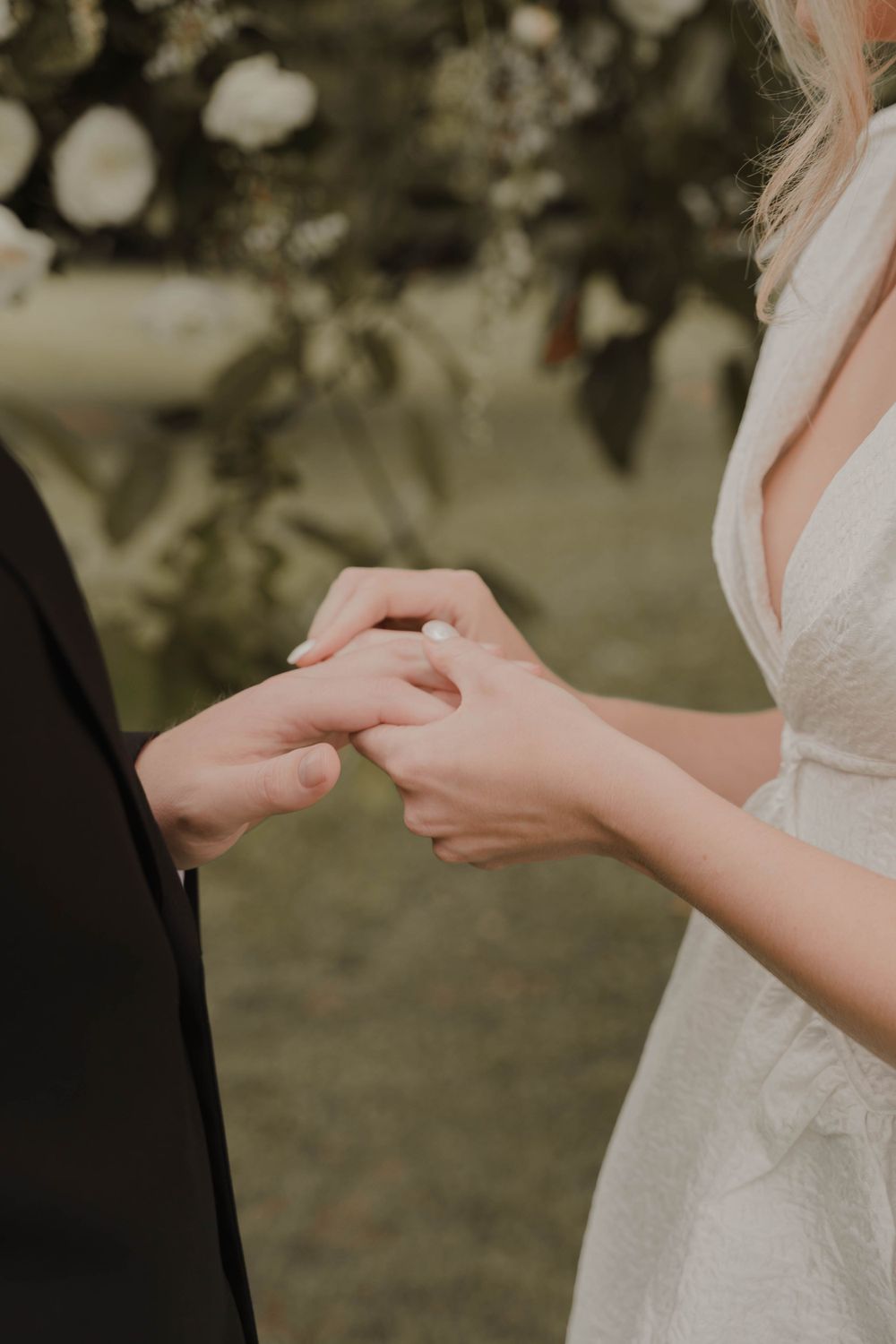 Close-up of couple exchanging wedding rings during outdoor ceremony with white flowers in background.