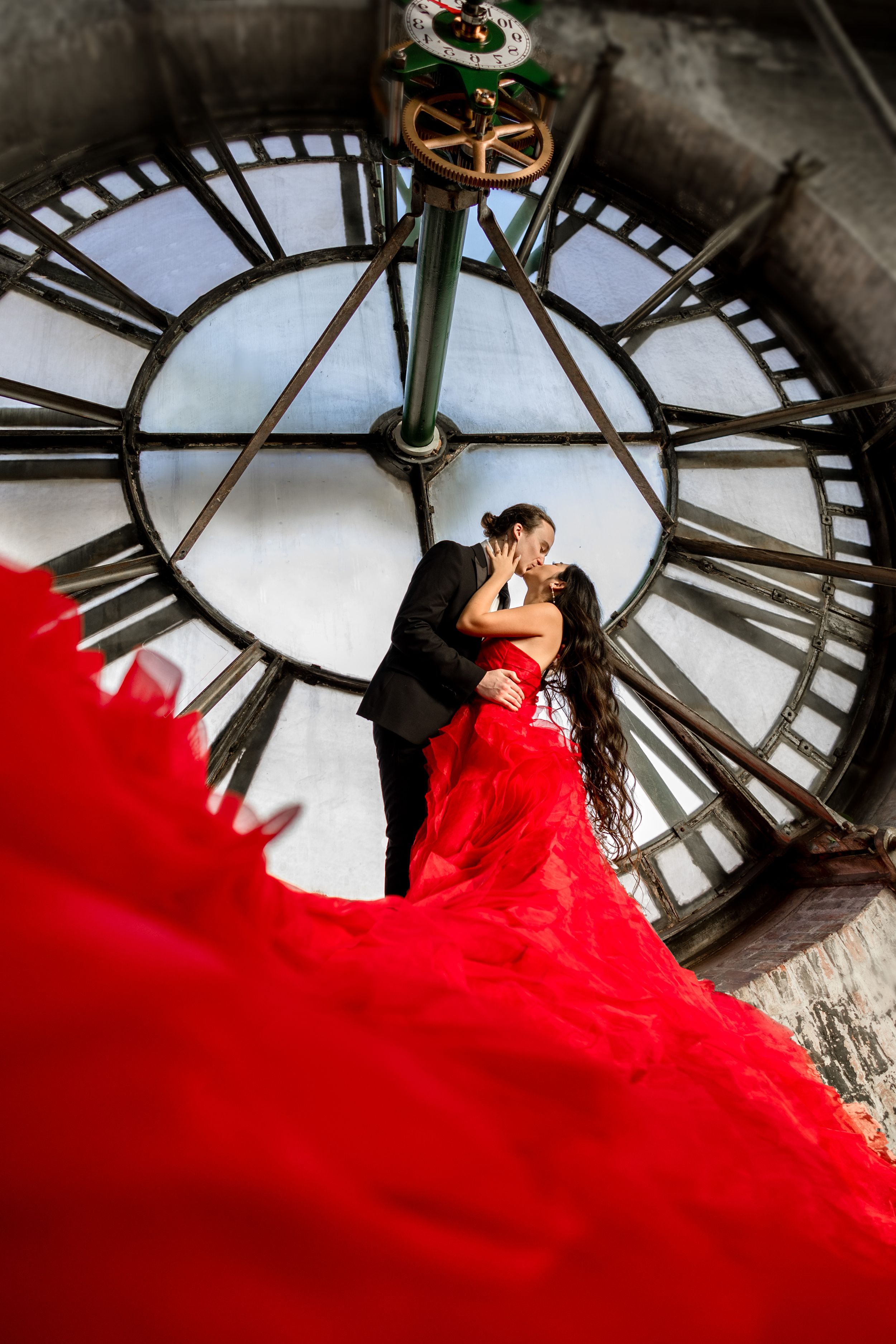 Romantic couple embraces inside Bromo Seltzer Arts Tower in front of large clock face with flowing red dress.