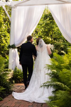 Newlyweds walk through a white draped wedding arch surrounded by lush garden ferns at the Elkridge Furnace Inn.