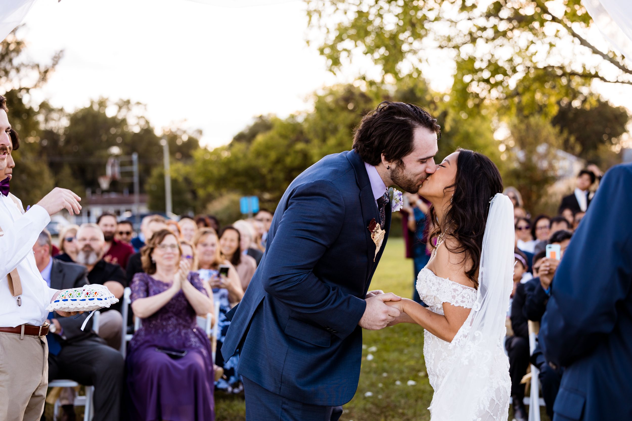 A joyous wedding ceremony kiss with guests clapping in the background on a sunny day.