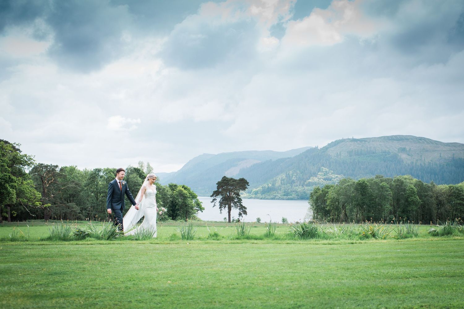 panoramic view of bride and groom walking across a lawn with Lake District mountains in the background