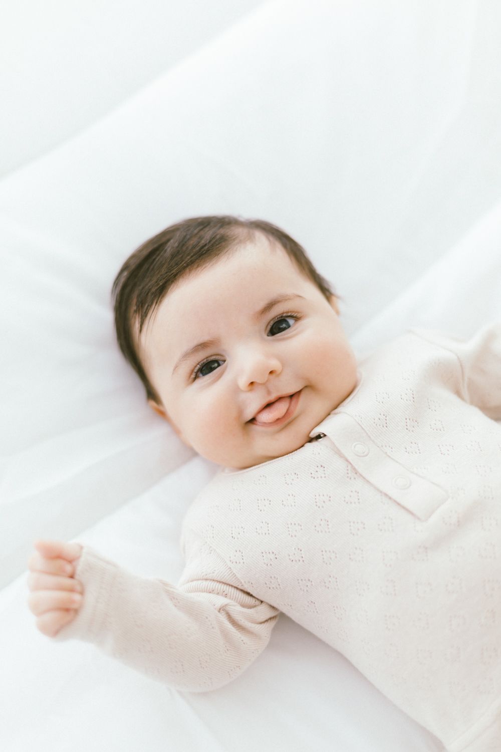 Smiling baby in light colored clothing rests on white blanket looking upward.