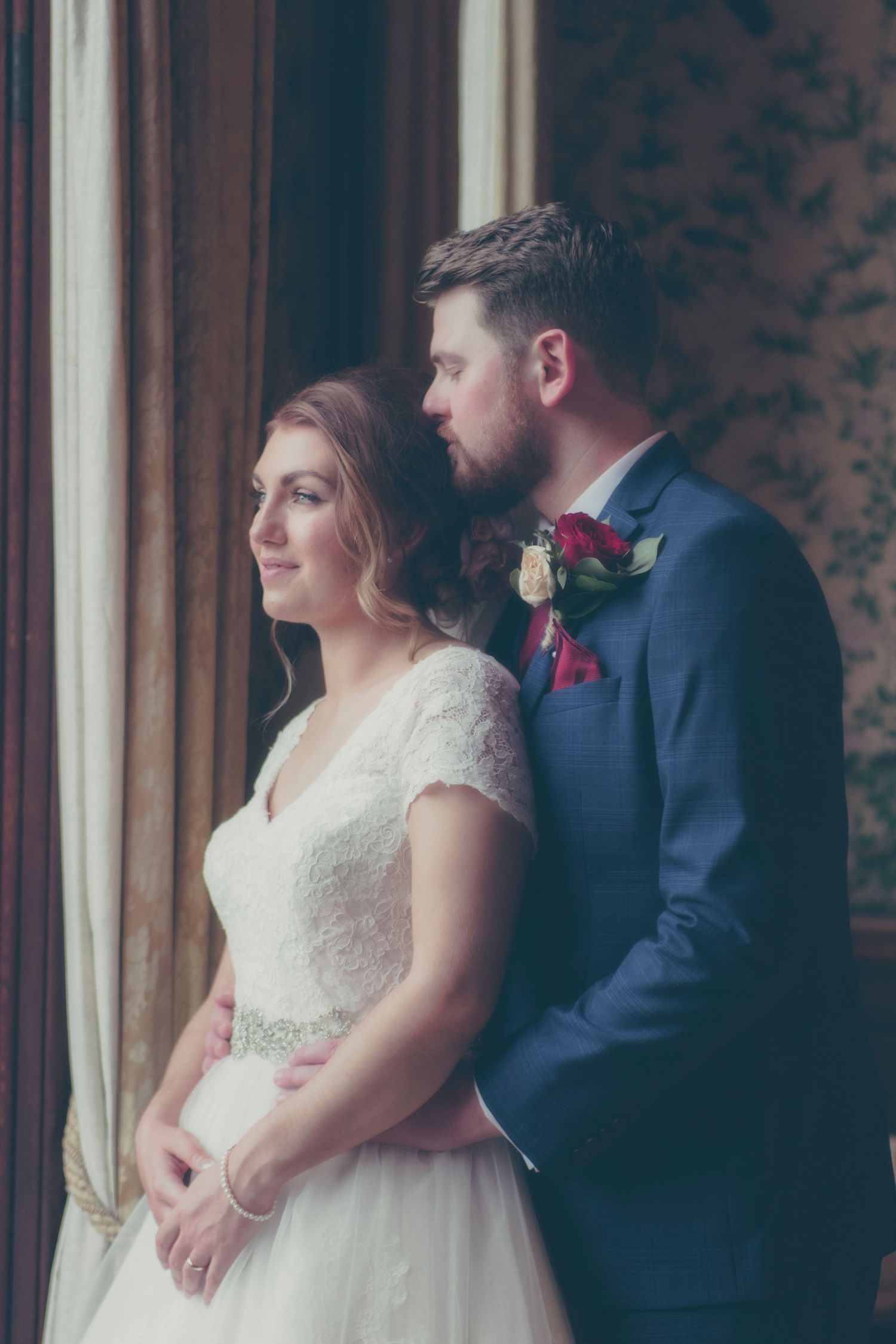 A couple stands together by a window in elegant wedding attire featuring a lace dress and navy suit.