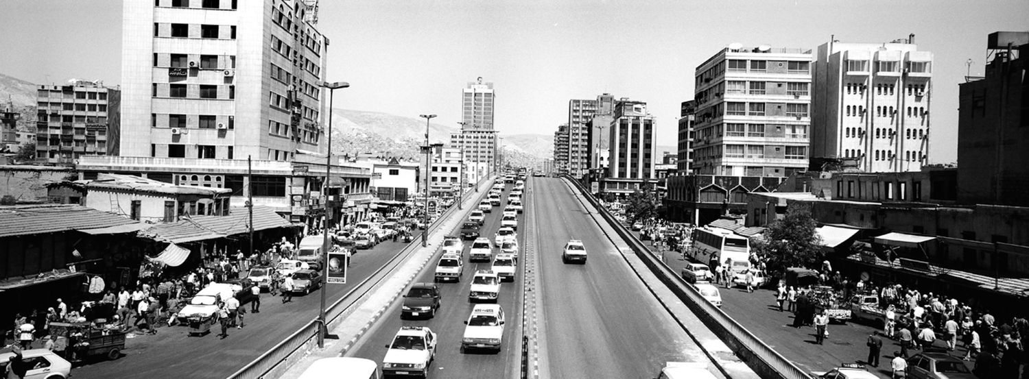 Vintage black and white cityscape showing a wide urban street with cars and buildings in what appears to be 1960s era.
