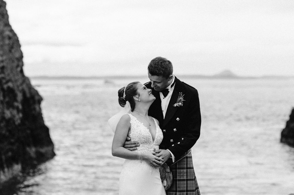 A romantic wedding moment captured against a misty ocean backdrop with traditional Scottish attire.