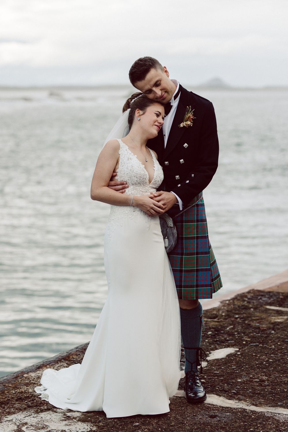 A tender embrace between newlyweds on a rocky shoreline with traditional Scottish wedding attire.