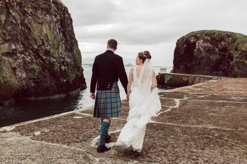 A couple walks along a dramatic rocky coastline during a wedding photoshoot in Scotland.