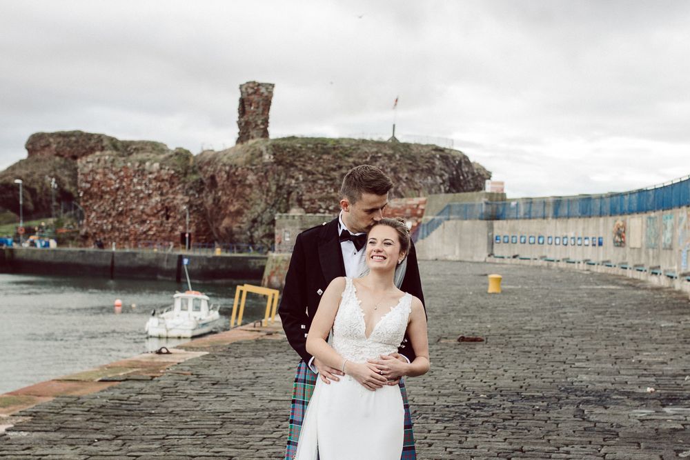 Wedding couple posing on stone pier with harbor and boats in background.