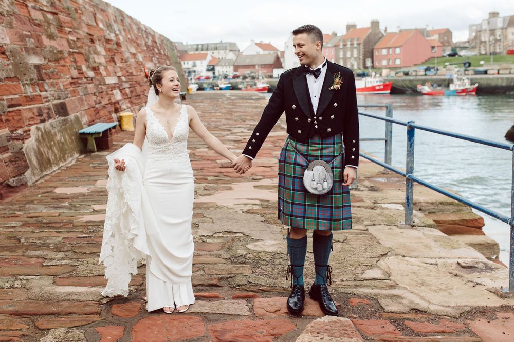 Newlyweds pose along historic brick walls near a harbor in traditional Scottish wedding attire.