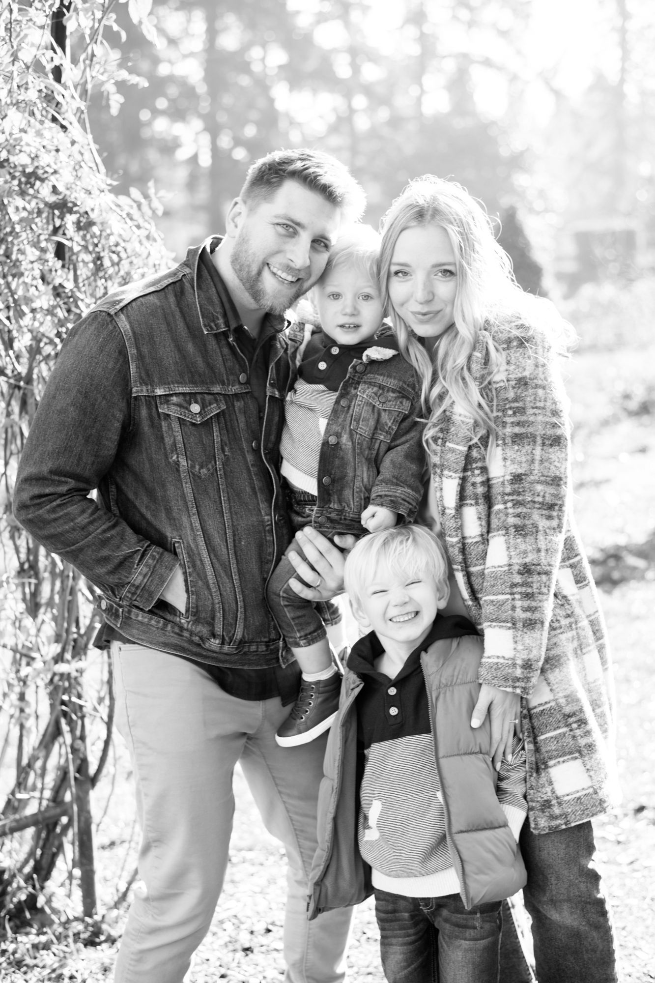 Black and white outdoor family portrait against tree in casual winter wear.