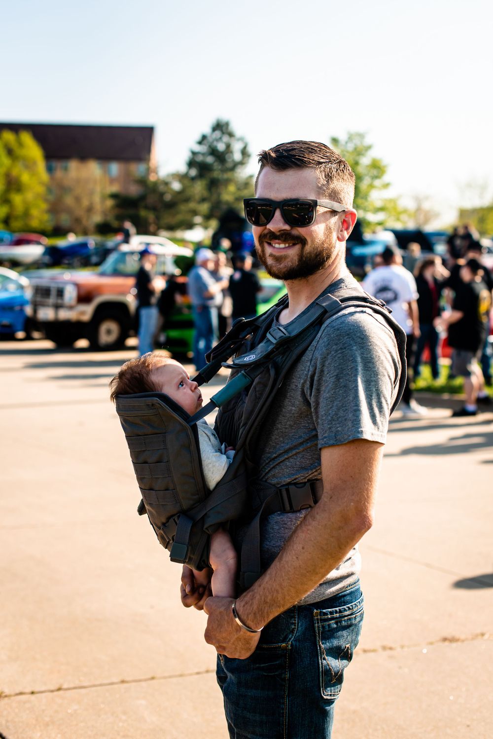 Parent wearing sunglasses carries baby in carrier at outdoor gathering with cars.
