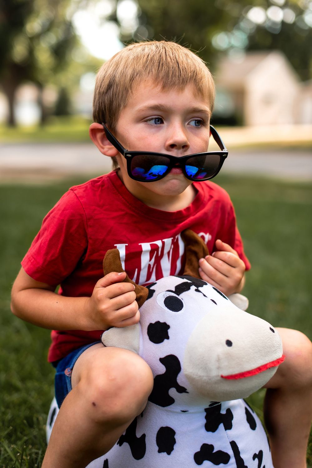 Child sitting on spotted cow toy wearing sunglasses in suburban setting.