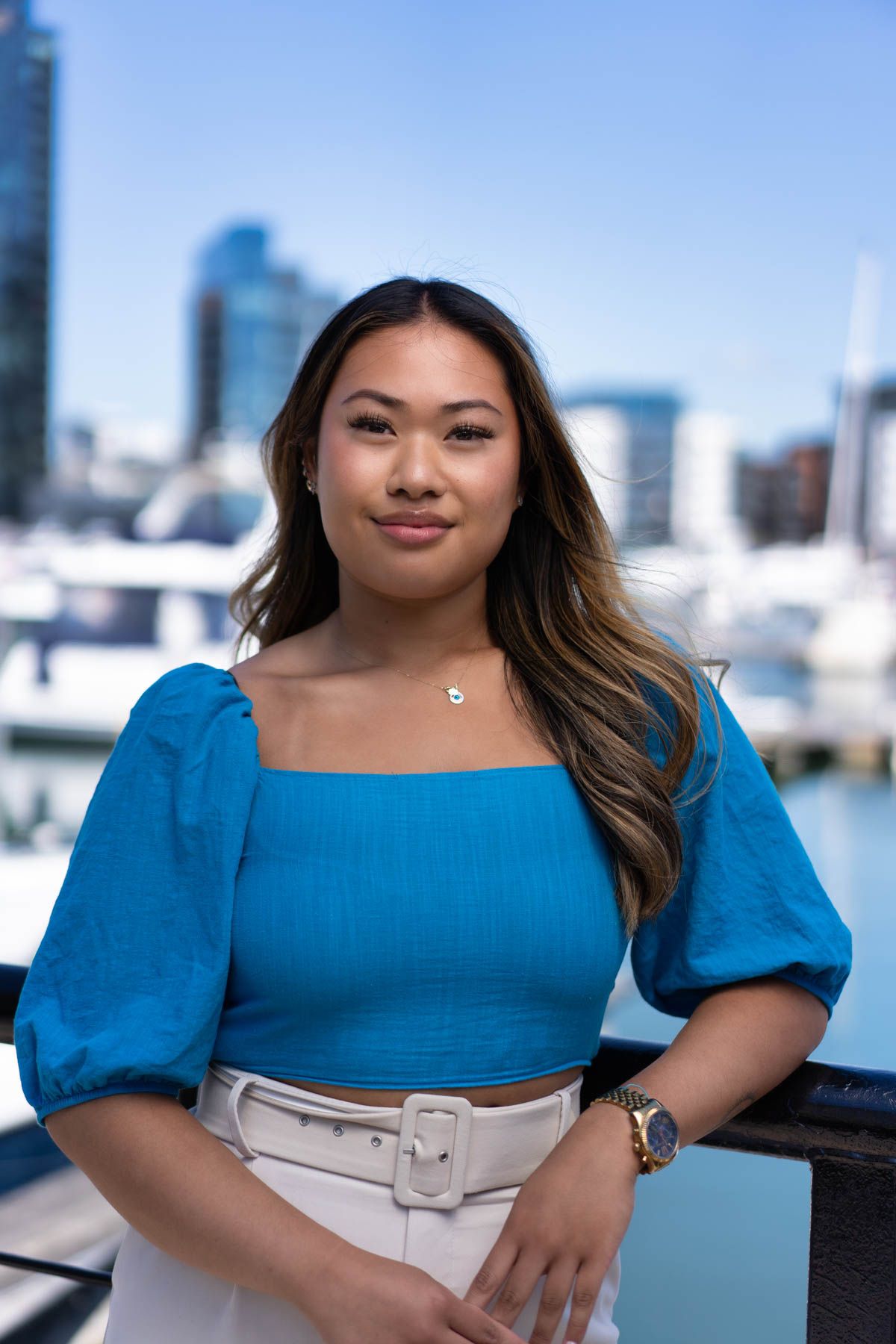Woman in blue top poses against waterfront backdrop with boats and city skyline.