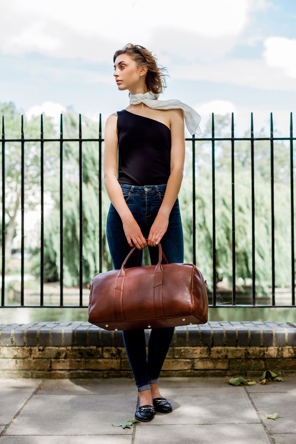 Stylish outfit featuring black top and brown leather duffle bag against iron fence.
