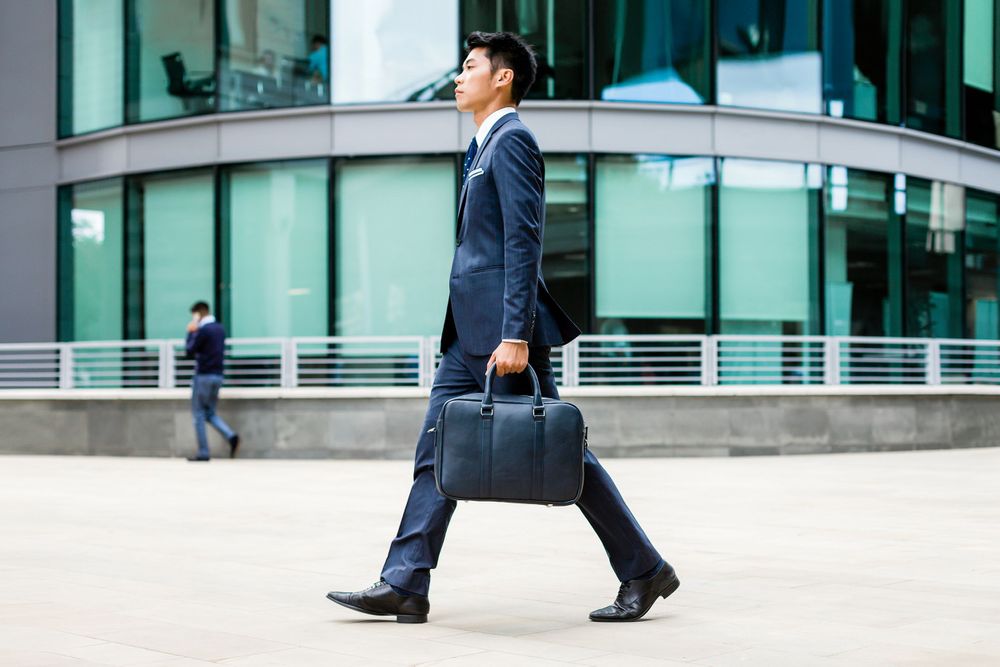 Business professional walking with navy briefcase near modern glass building.