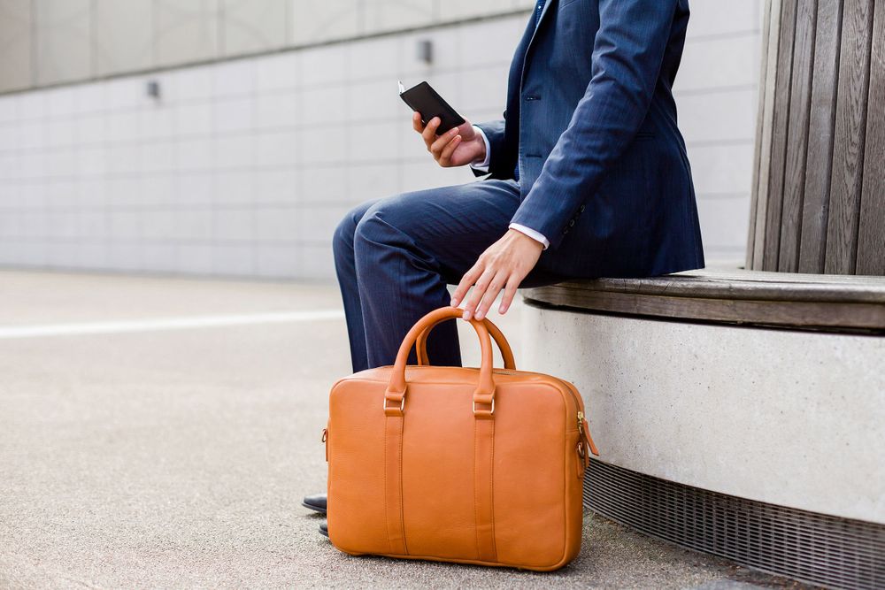 Person in business wear using phone with tan leather briefcase on bench.