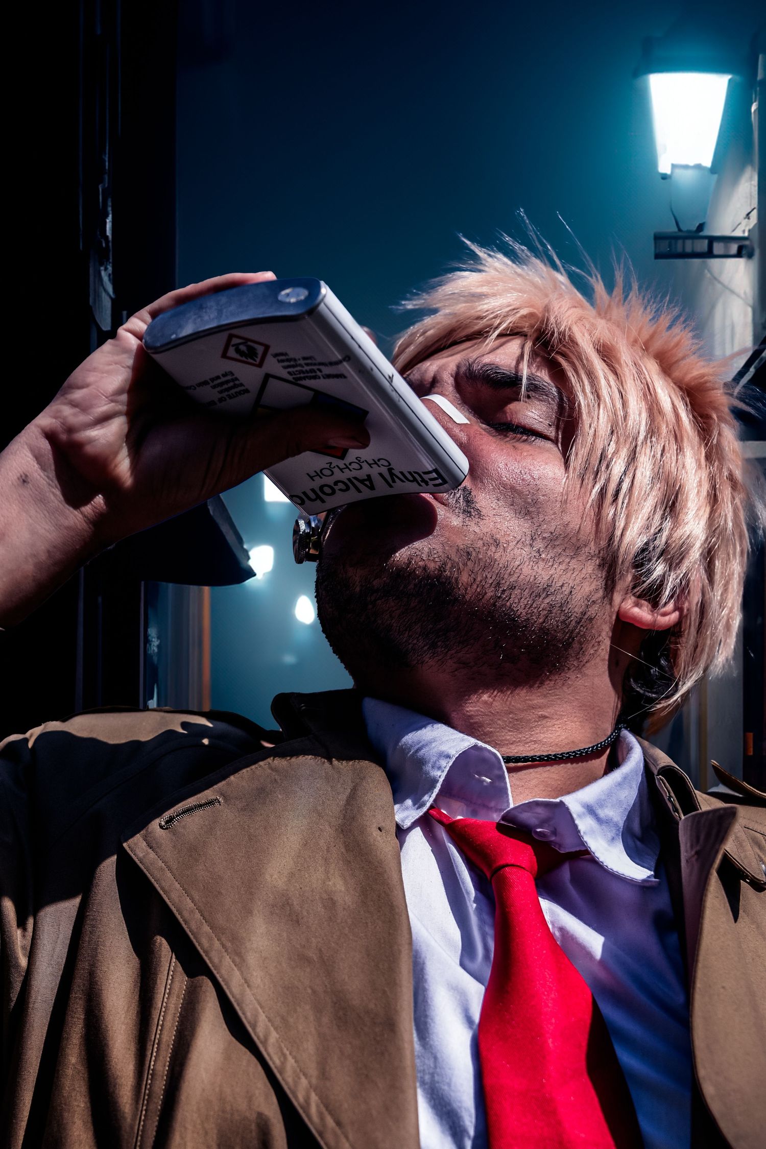 Person in brown coat and red tie drinking from a flask against a dark background with street lamp.