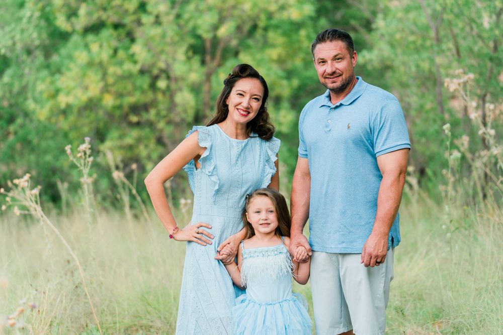 A family in coordinating blue outfits poses together in a sunny meadow setting.