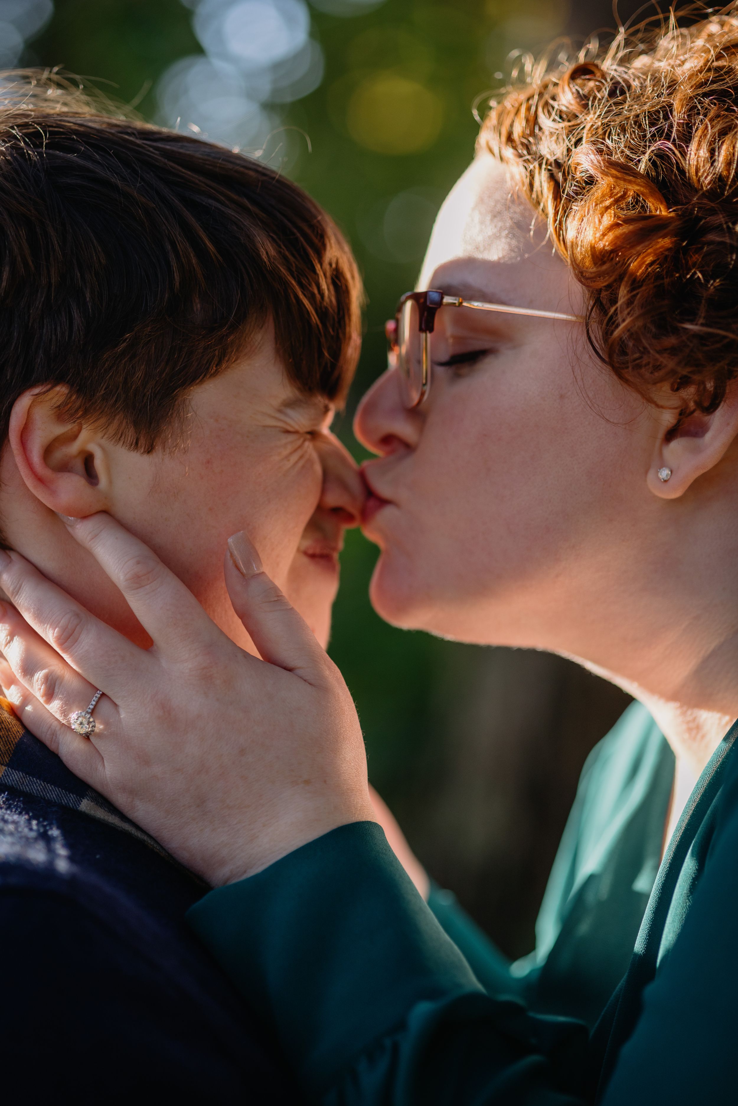 Two women share a tender kiss outdoors in warm golden sunlight during an engagement session.