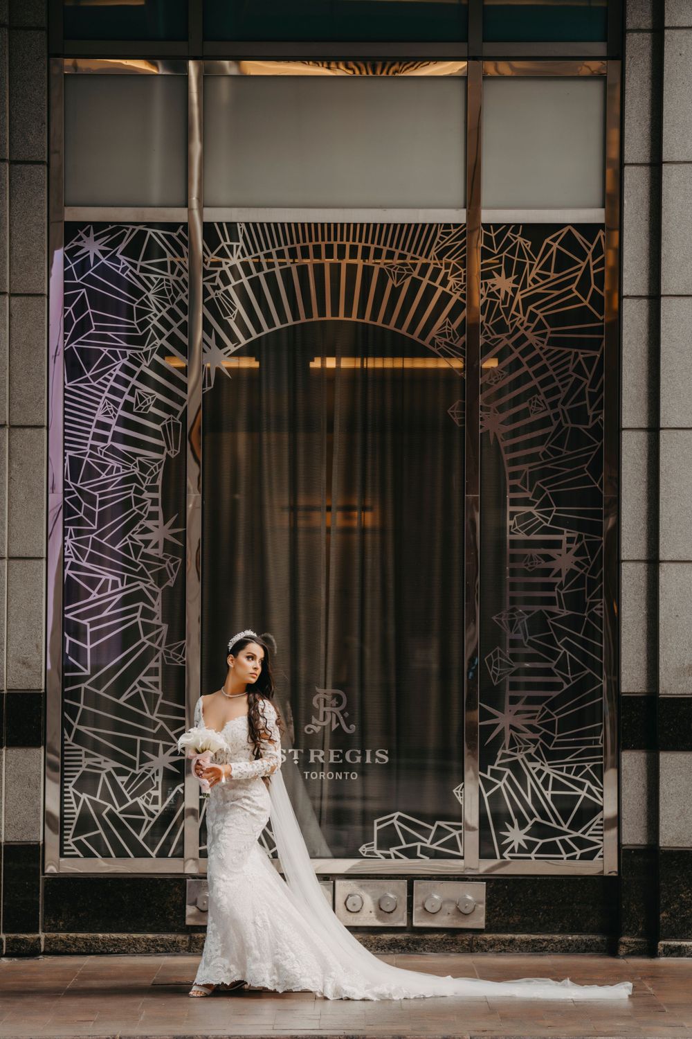 Bride in fitted lace wedding gown poses elegantly in front of St. Regis hotel entrance with decorative metalwork.