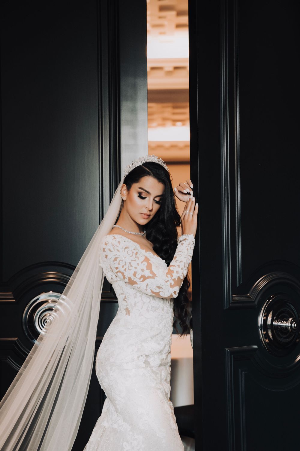Elegant bride in lace long-sleeved wedding dress poses dramatically against dark paneled doorway.