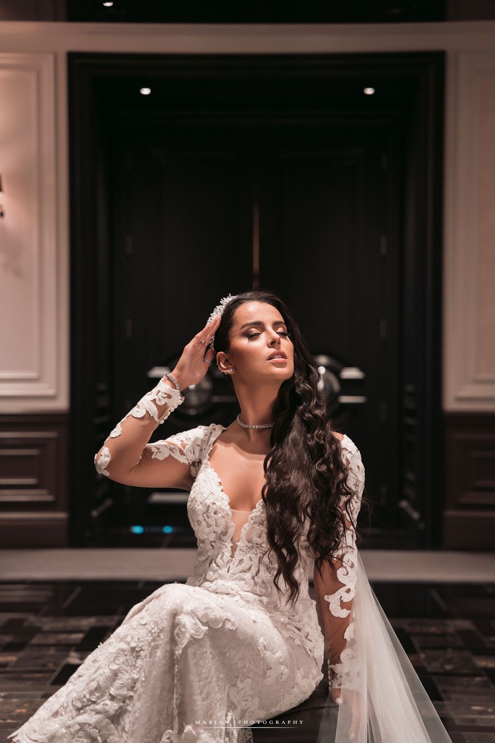 Bride in long-sleeved lace gown poses dramatically in elegant indoor setting.