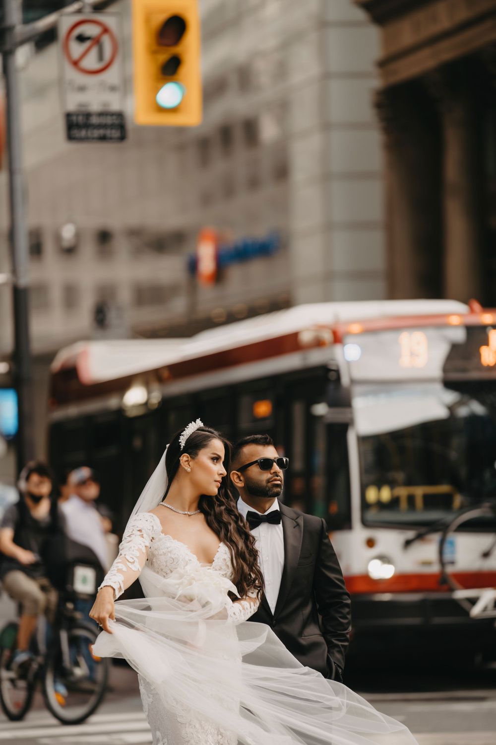 Newlywed couple in formal attire walking through city streets with flowing white wedding dress and TTC bus in background.