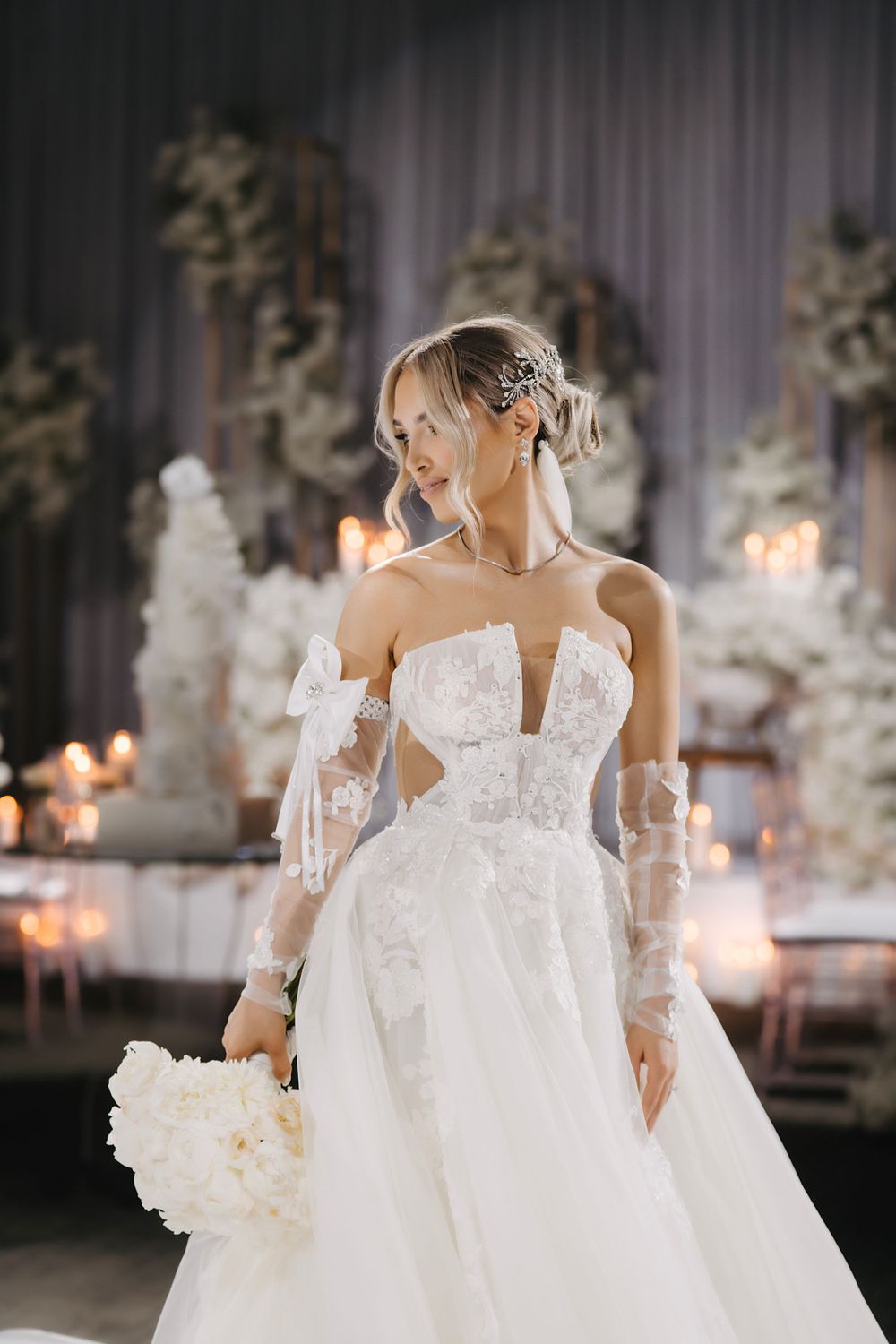 Bride wearing an elegant white gown with lace sleeves stands in a candlelit reception venue.