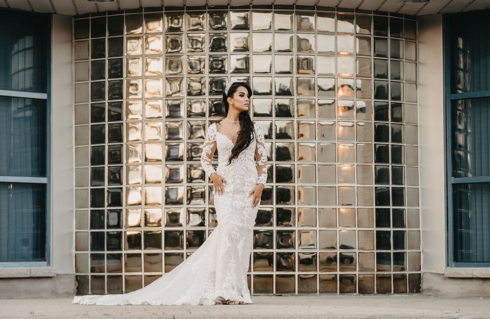 Bride in fitted lace wedding dress poses against modern glass block wall during urban photoshoot.