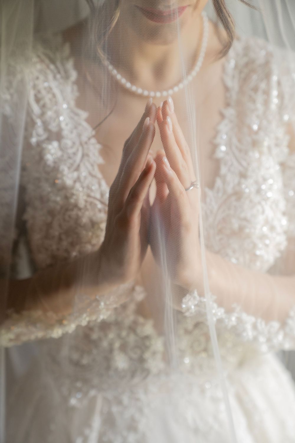 Close-up of bride's praying hands with pearl necklace and lace wedding dress details.