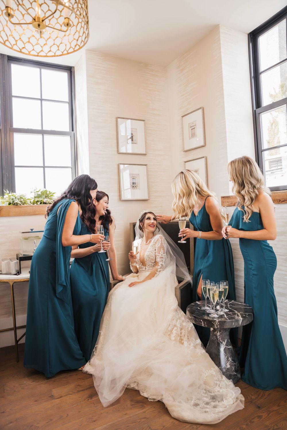 Bride in white gown surrounded by bridesmaids in teal dresses sharing champagne and laughter.