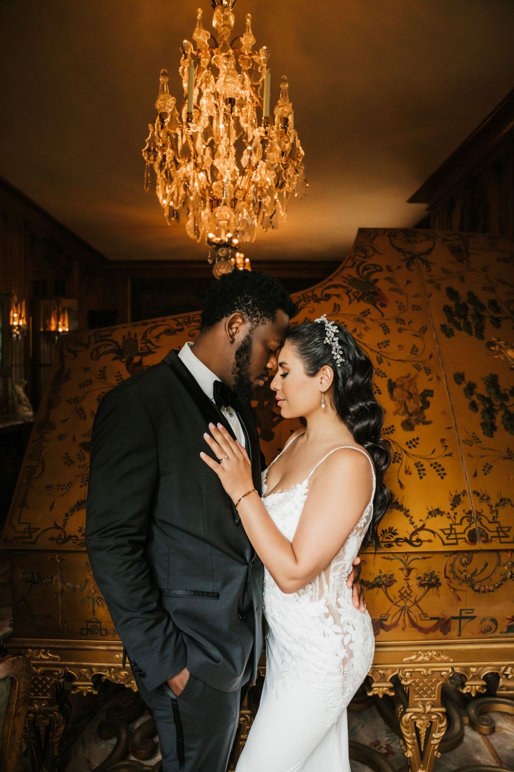 Wedding couple poses intimately beneath an ornate crystal chandelier in an elegant room.