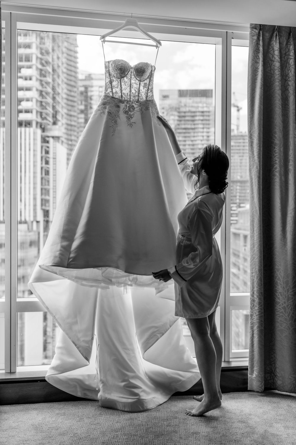 Wedding dress hanging by window while bride in robe admires it.