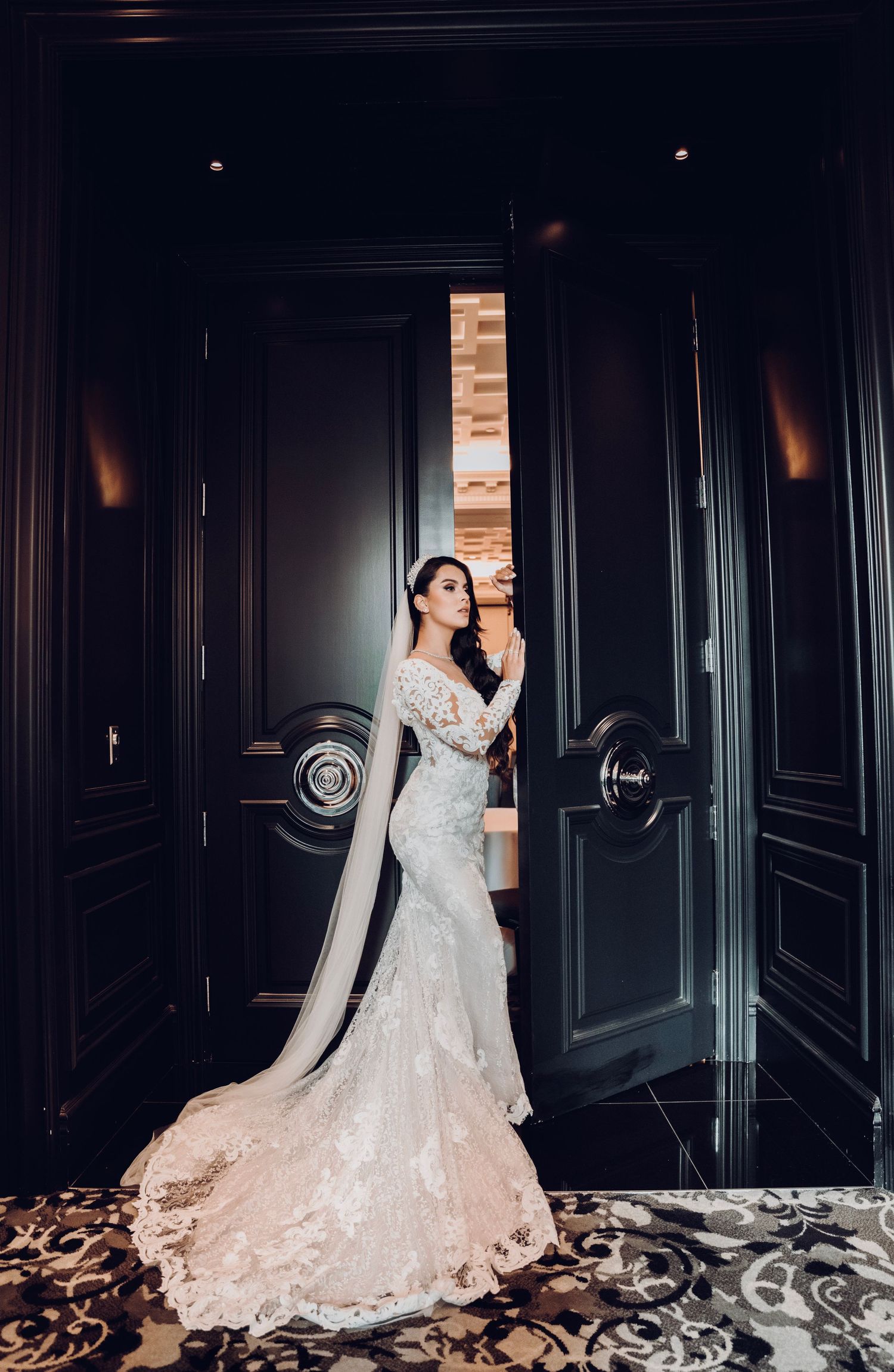 Dramatic portrait of bride in fitted lace gown with cathedral veil against dark doorway.