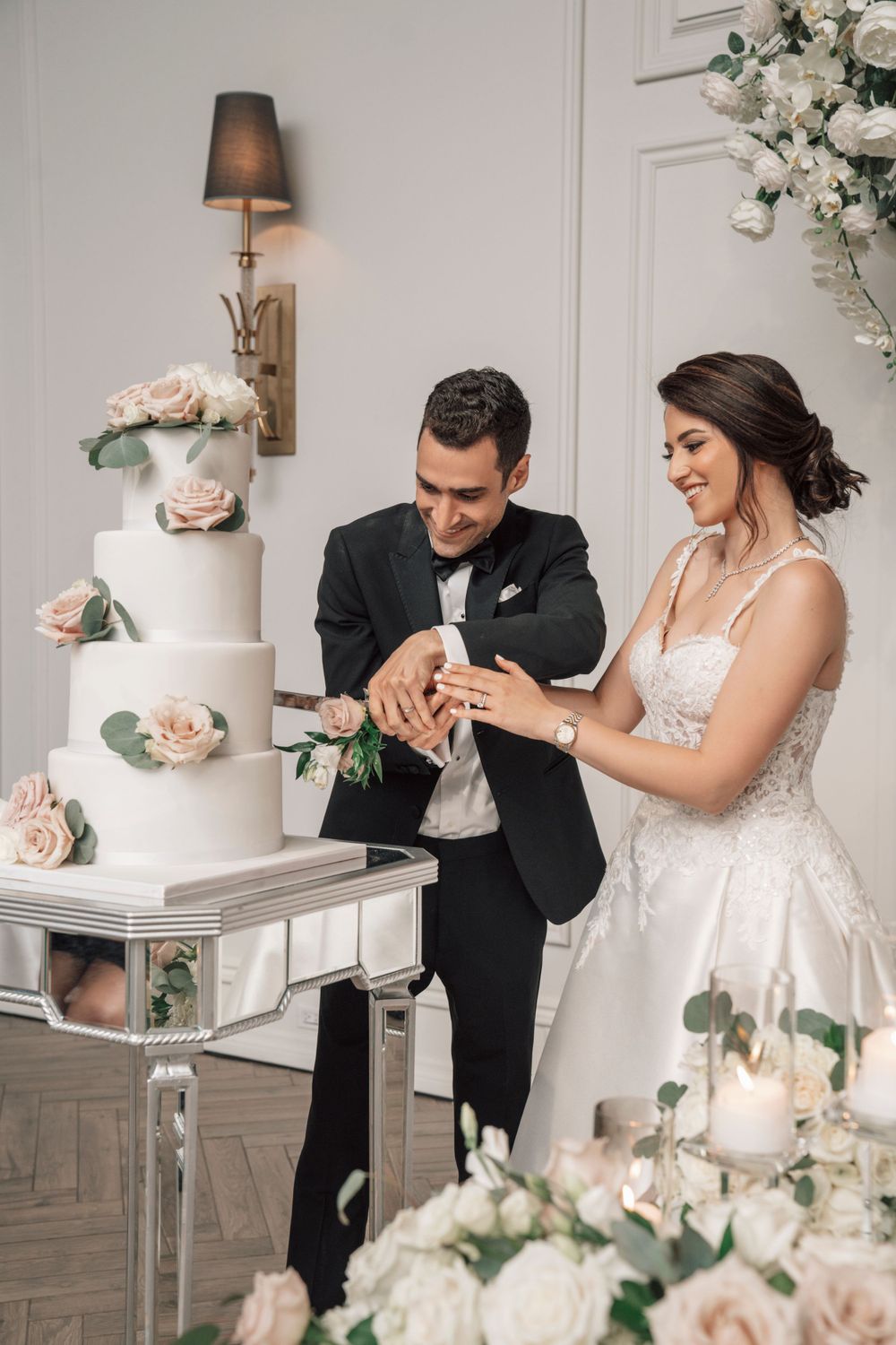 Newlyweds cutting elegant white tiered wedding cake decorated with blush roses.