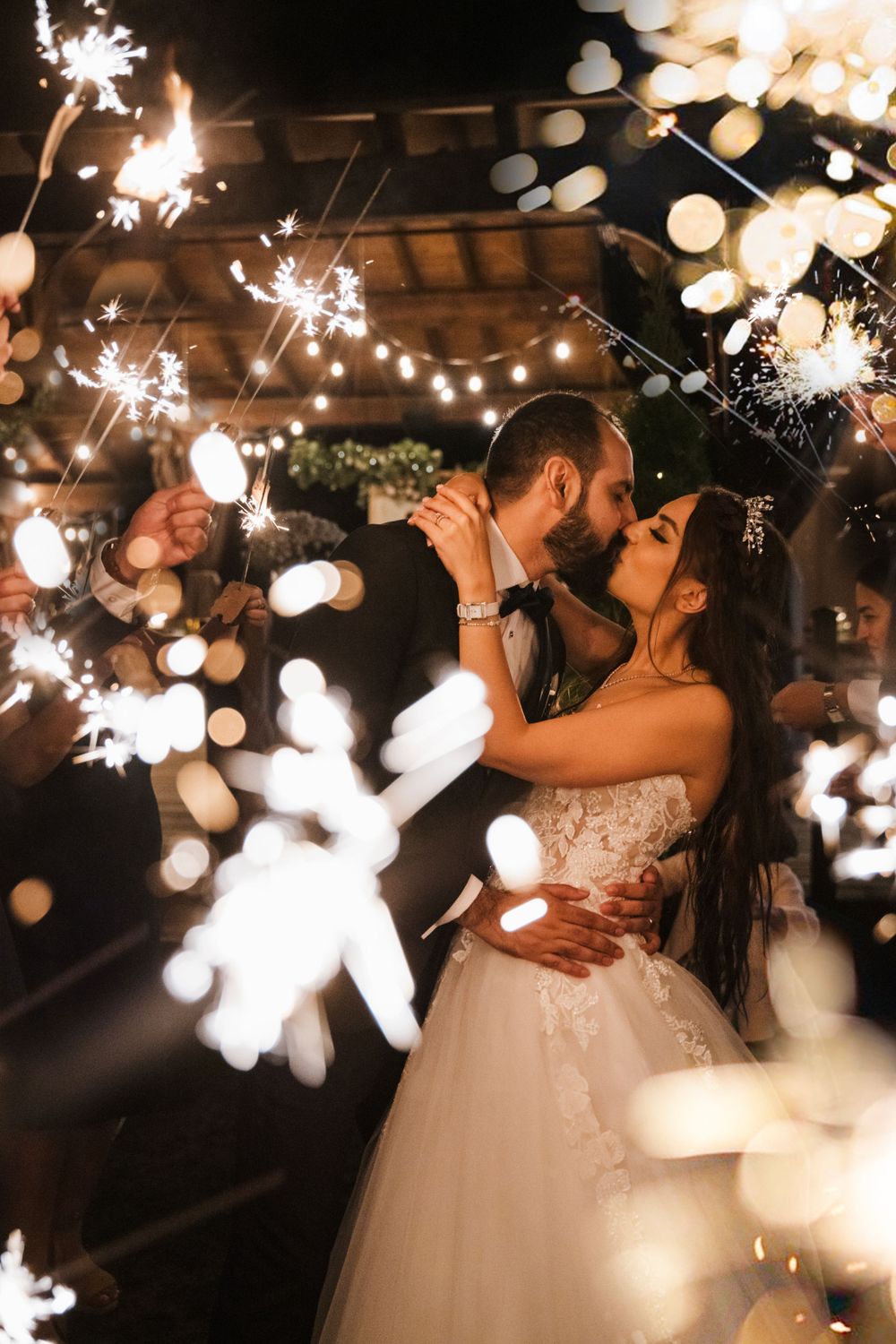Newlyweds share a romantic kiss under twinkling lights and sparklers at their evening wedding celebration.