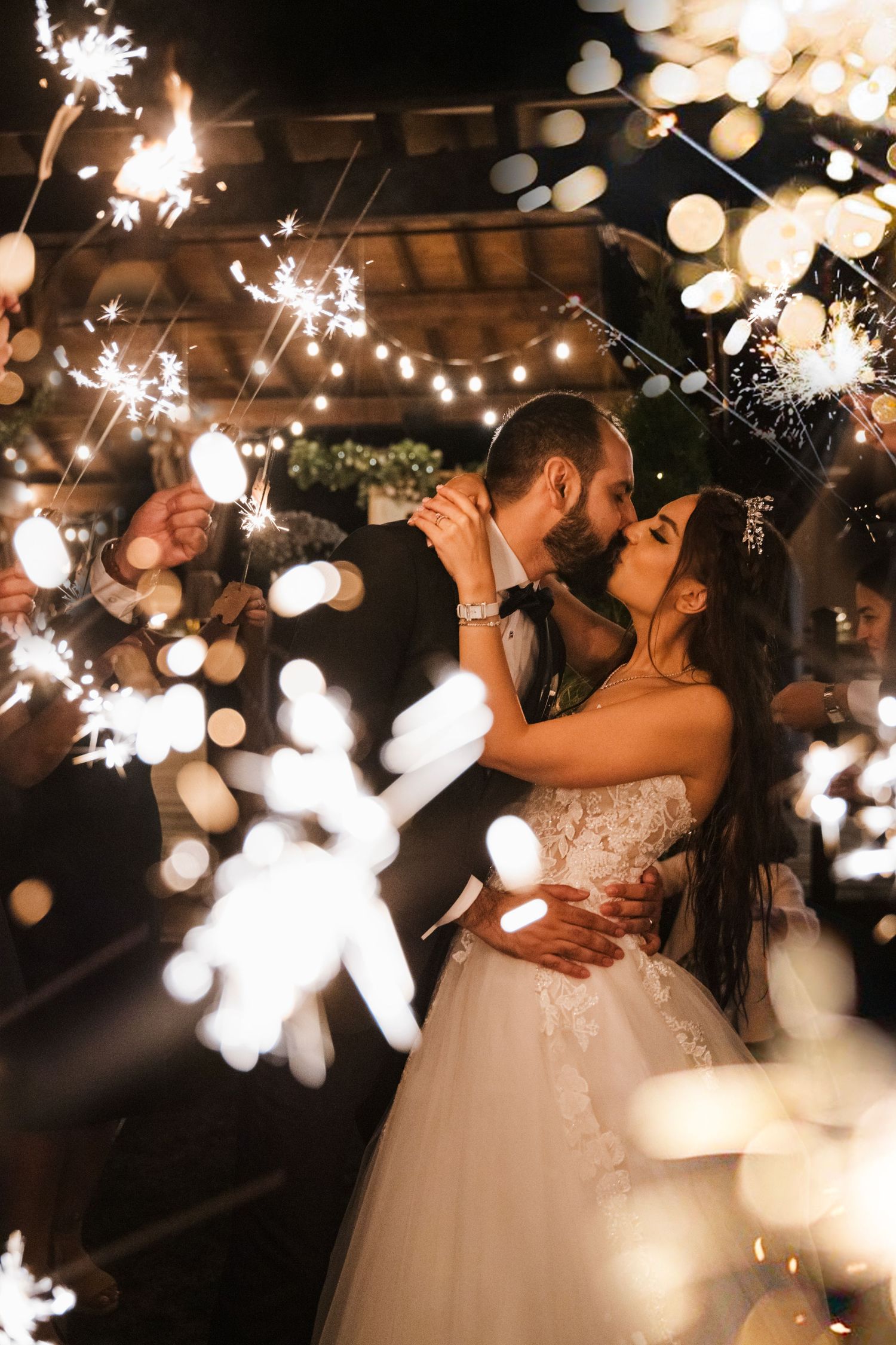 Newlyweds share a romantic kiss under twinkling lights and sparklers at their evening wedding celebration.