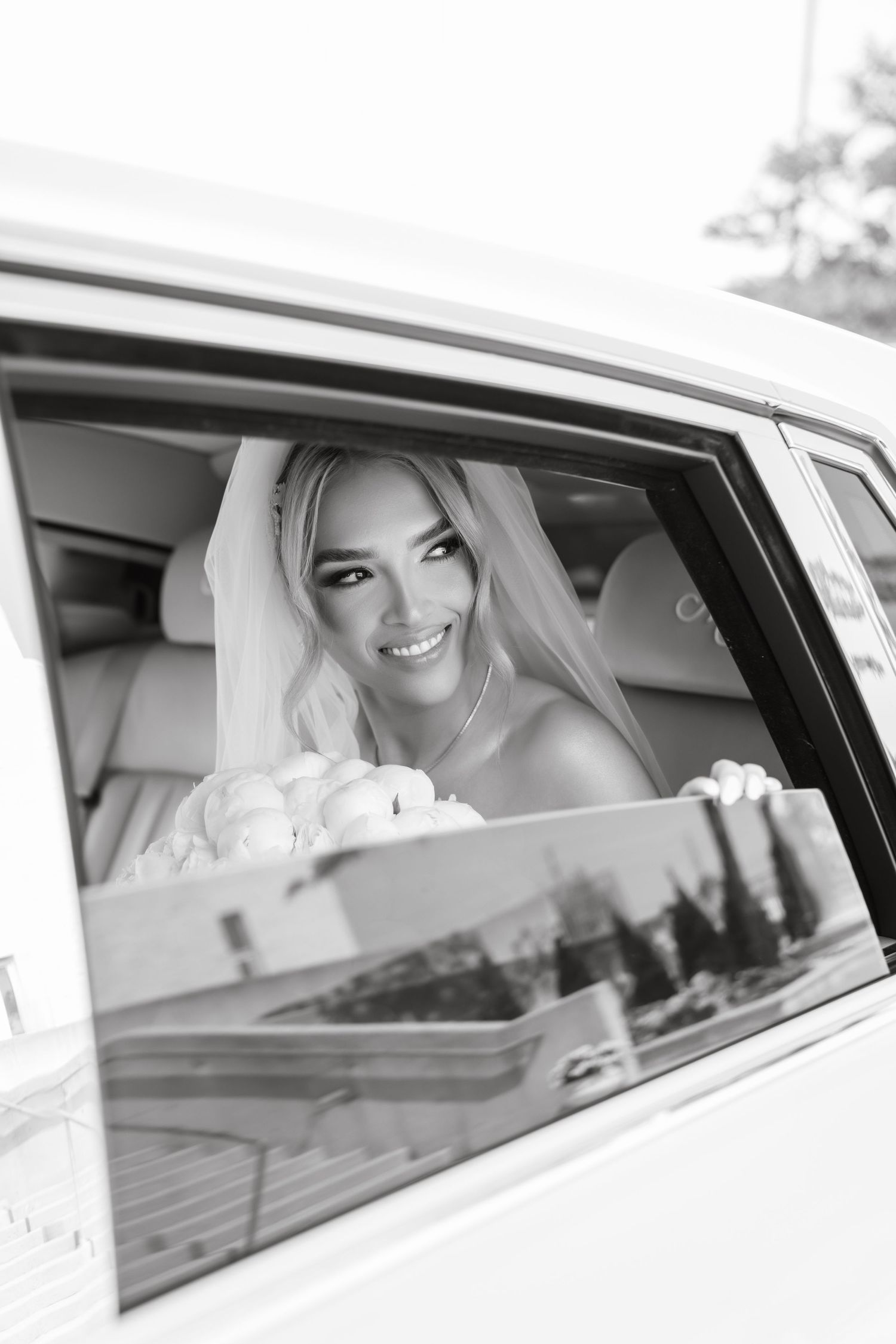 A joyful bride smiles while sitting in a luxury car window.