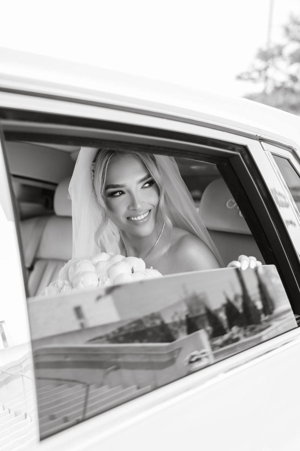 A joyful bride smiles while sitting in a luxury car window.