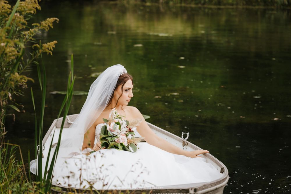 A bride in a white dress sits gracefully by a tranquil pond while holding a wedding bouquet.