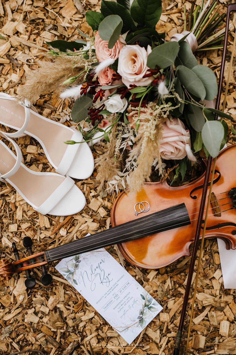 Flatlay of wedding details including white shoes, violin, roses and invitation on wooden background.