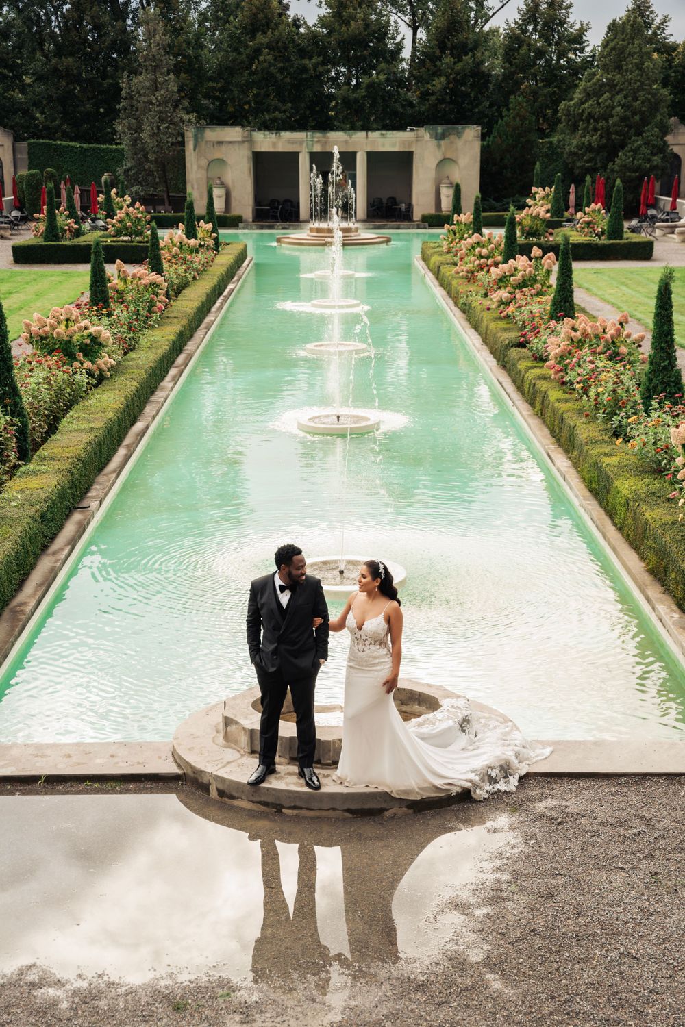 Wedding couple stands on a fountain platform surrounded by a manicured garden with water features.