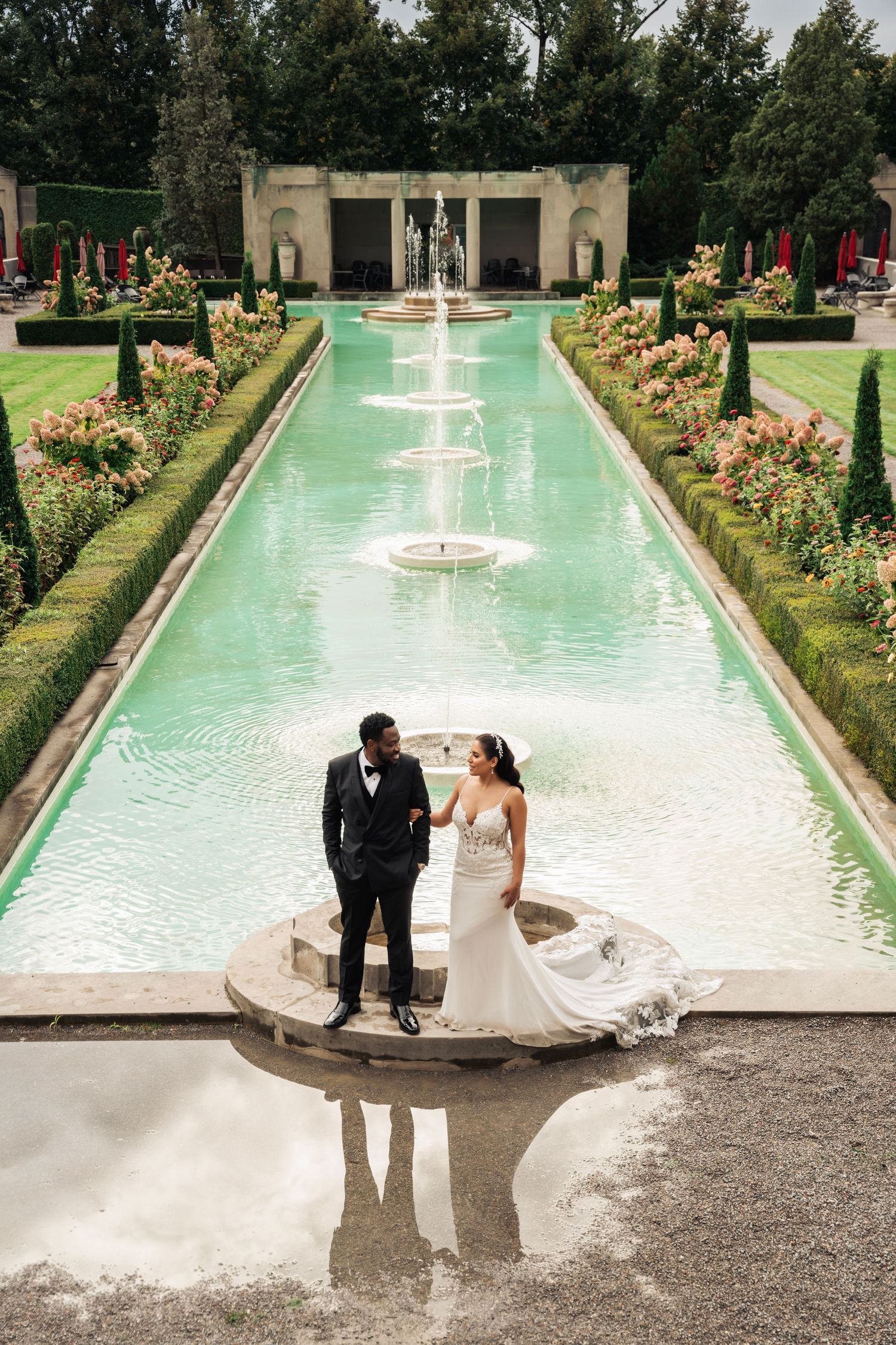 Wedding couple stands on a fountain platform surrounded by a manicured garden with water features.