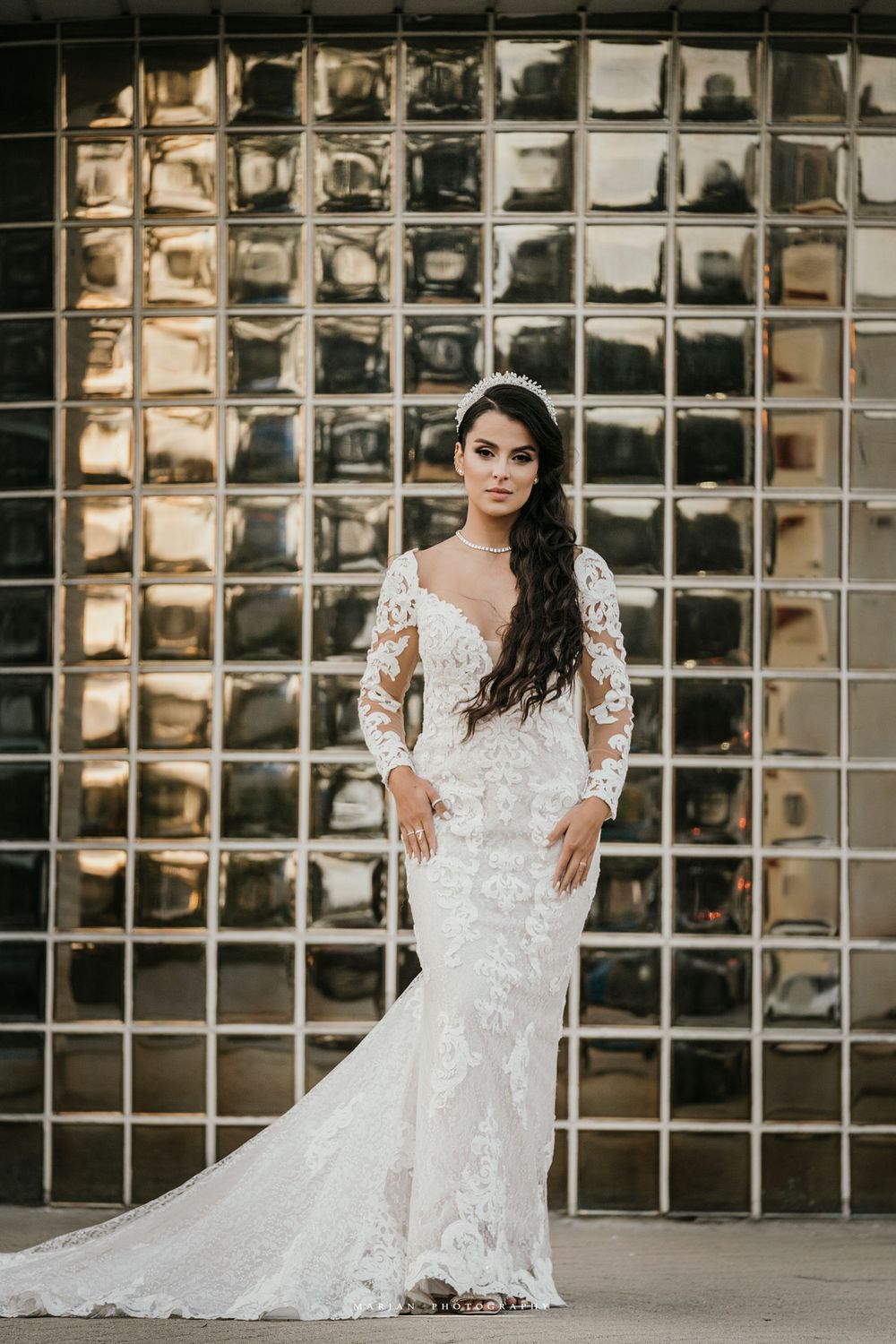 Bride in fitted lace wedding gown poses confidently against metallic glass block wall background.