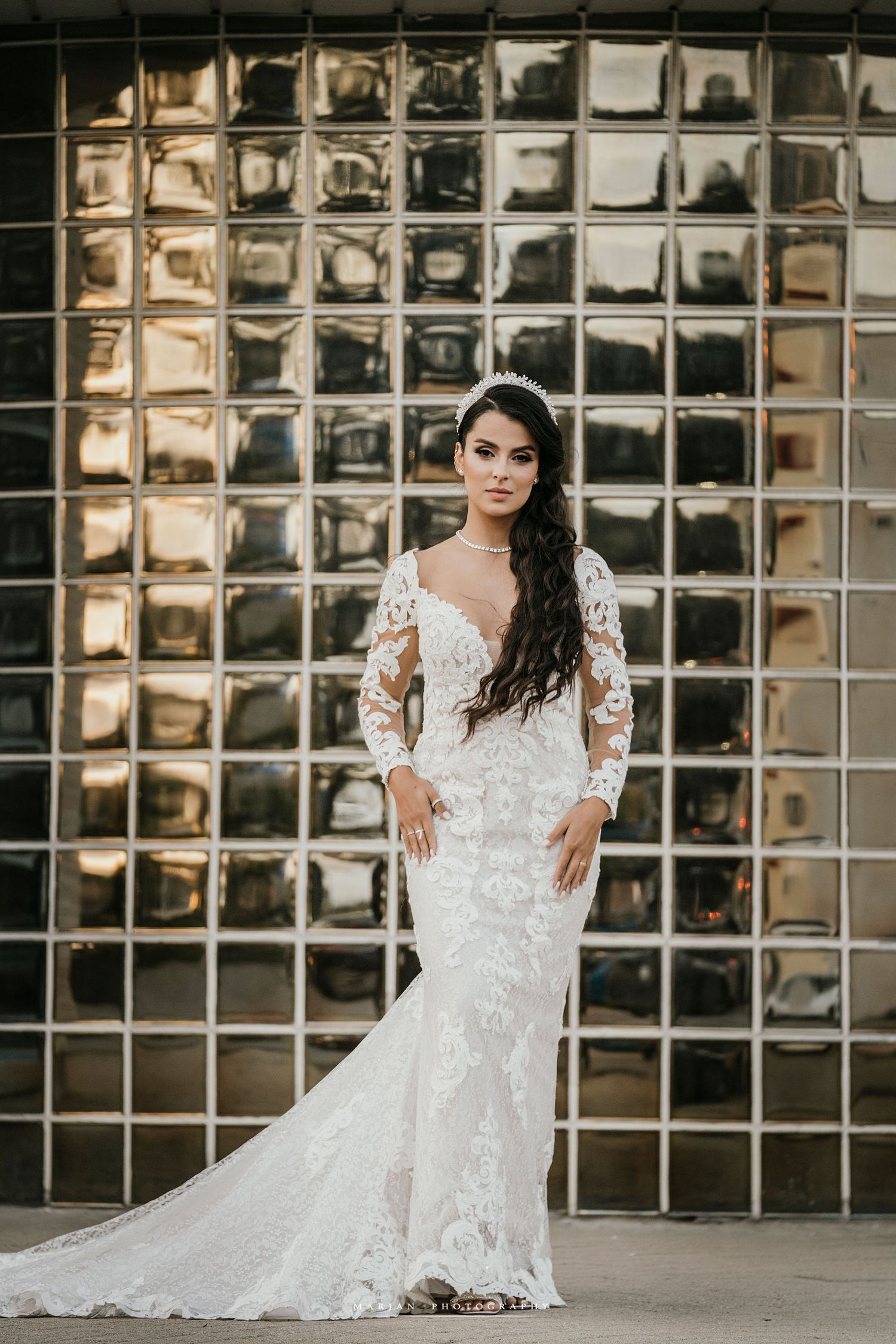 Bride in fitted lace wedding gown poses confidently against metallic glass block wall background.