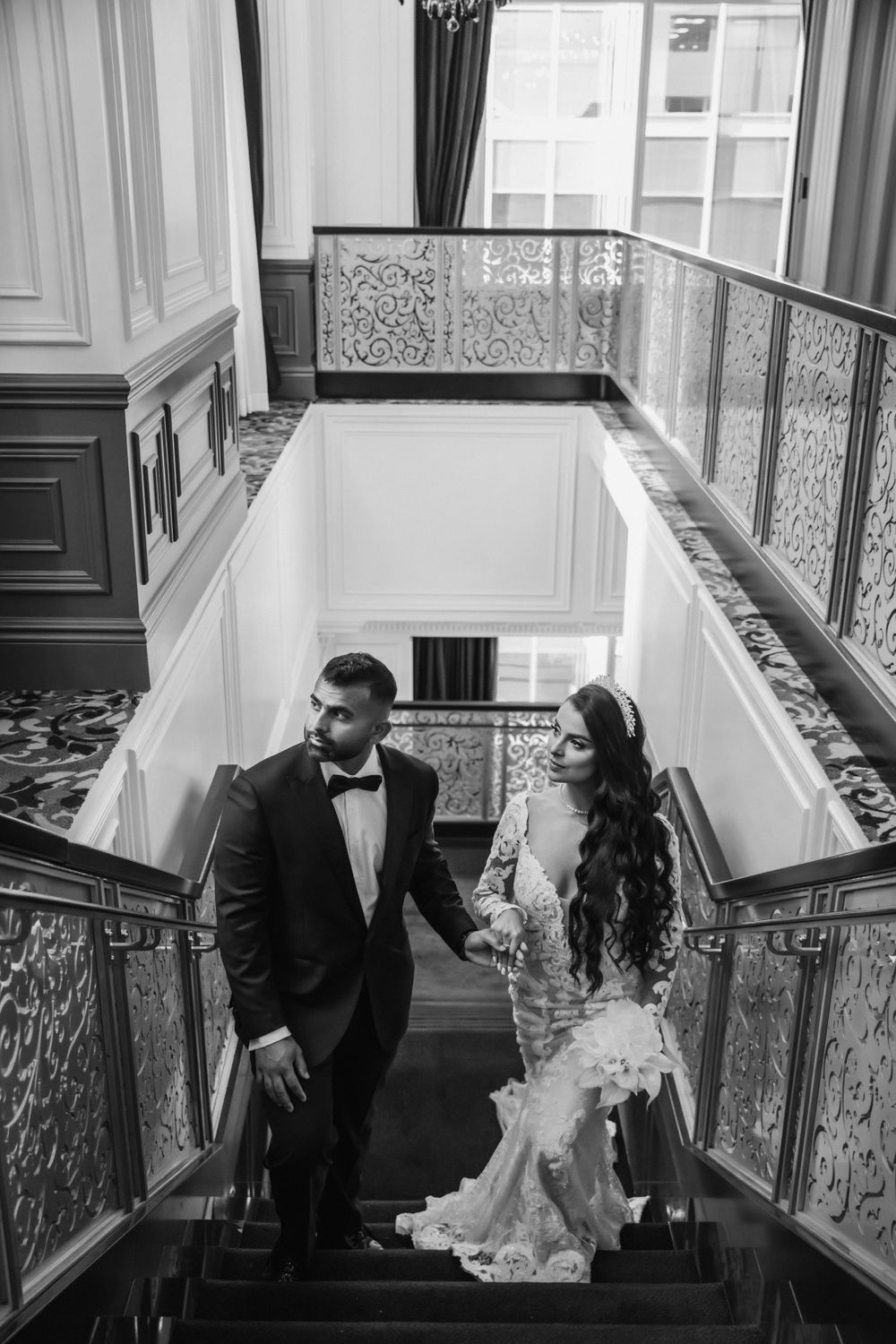 Black and white wedding photo of couple descending ornate staircase in formal attire.