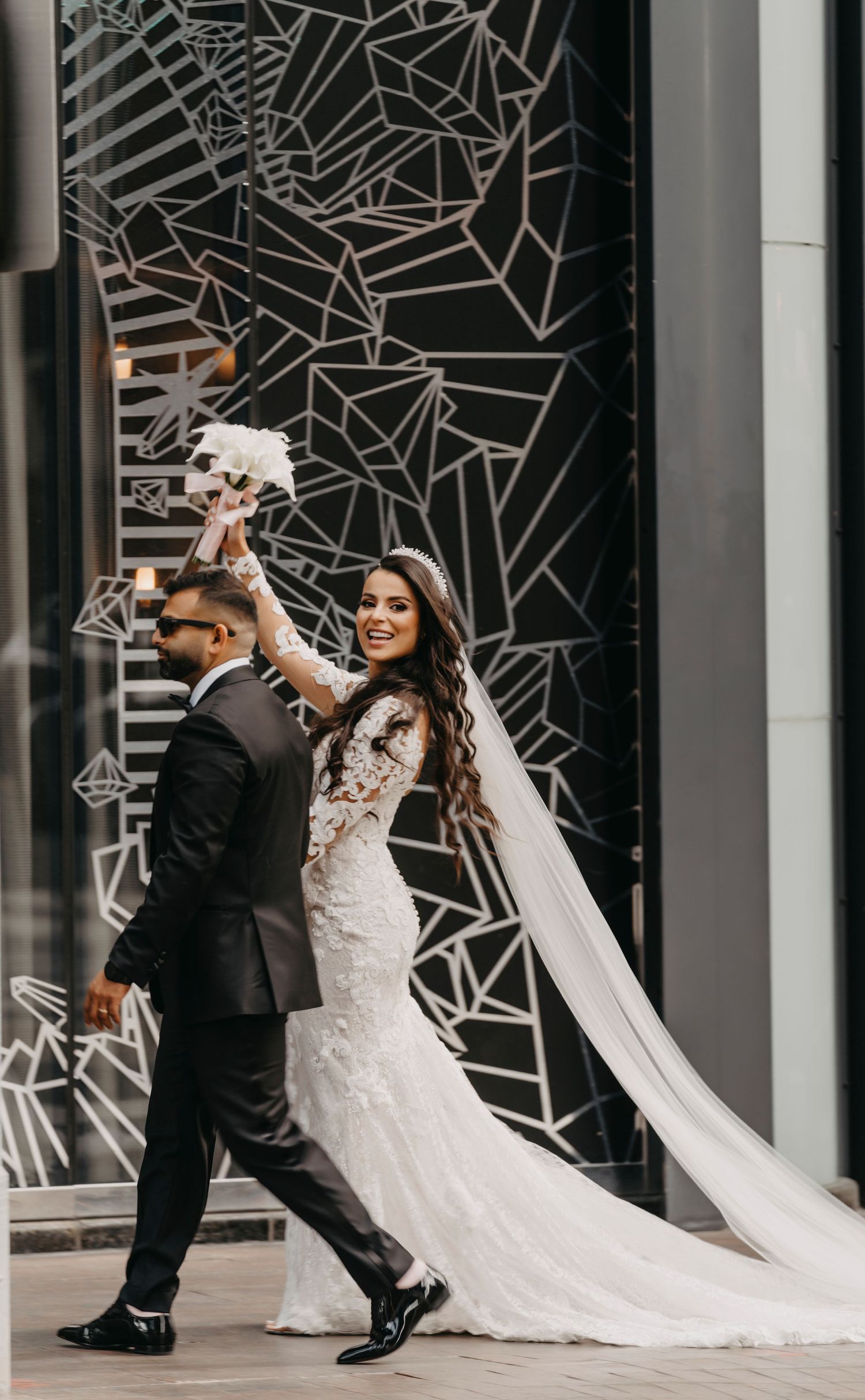 Bride in lace gown twirls with groom against geometric patterned wall during urban wedding photoshoot.