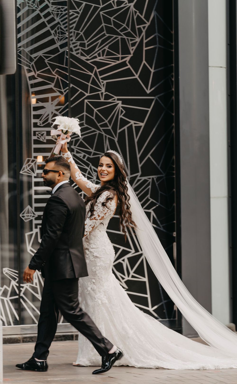 Bride in lace gown twirls with groom against geometric patterned wall during urban wedding photoshoot.